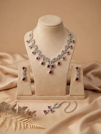 Silver-toned jewelry set displayed on beige mannequin stands, including a necklace, matching earrings, and a bracelet, each featuring red heart-shaped stones, set against a draped beige fabric background.